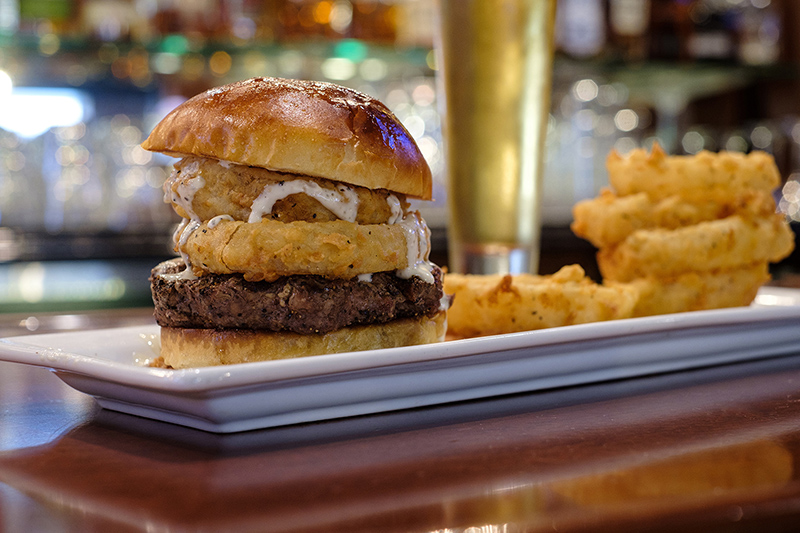 Picture of a hamburger served with onion rings and a glass of beer served at a bar at Bass Street Chop House